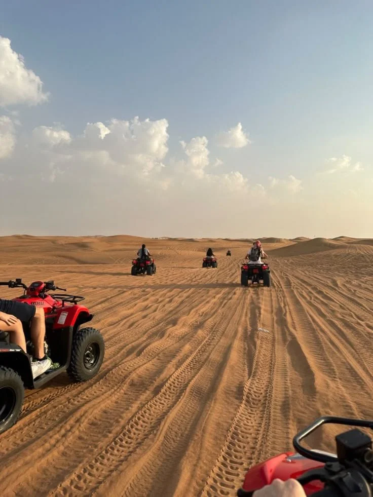 People riding all-terrain vehicles across sandy desert dunes under a partly cloudy sky.