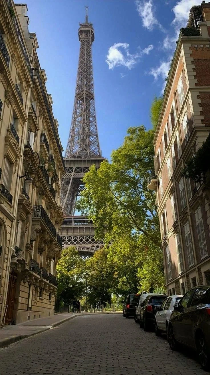 View of the Eiffel Tower from a city street with parked cars, buildings, trees, and a clear blue sky.
