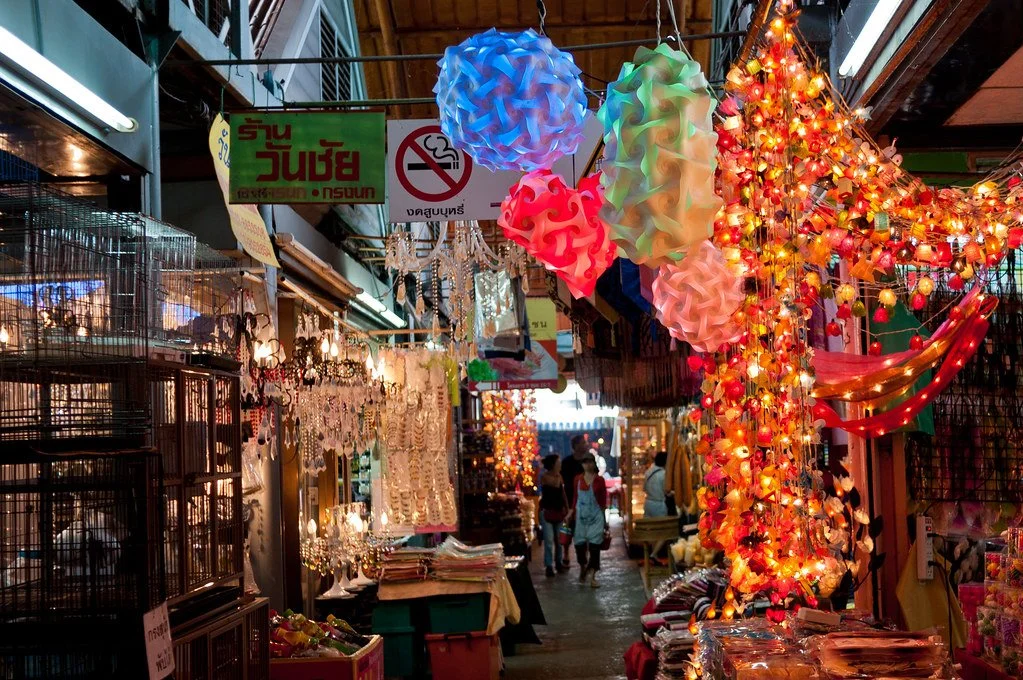 An indoor market with colorful hanging lanterns and lights, various stalls selling jewelry and decorative items, colorful tissue paper lanterns, and people shopping