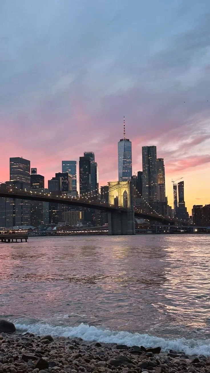 Sunset over the New York City skyline with Brooklyn Bridge in the foreground, reflecting on the water.