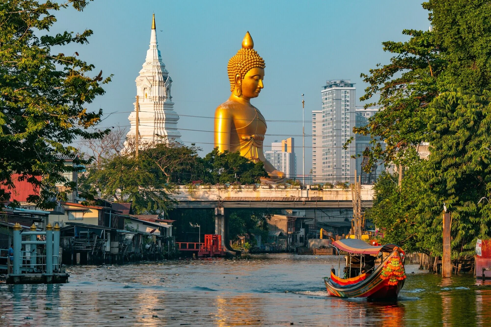 A river scene with a boat in the foreground, trees on the sides, and a large golden Buddha statue along with a white stupa in the background, with tall modern buildings behind them.