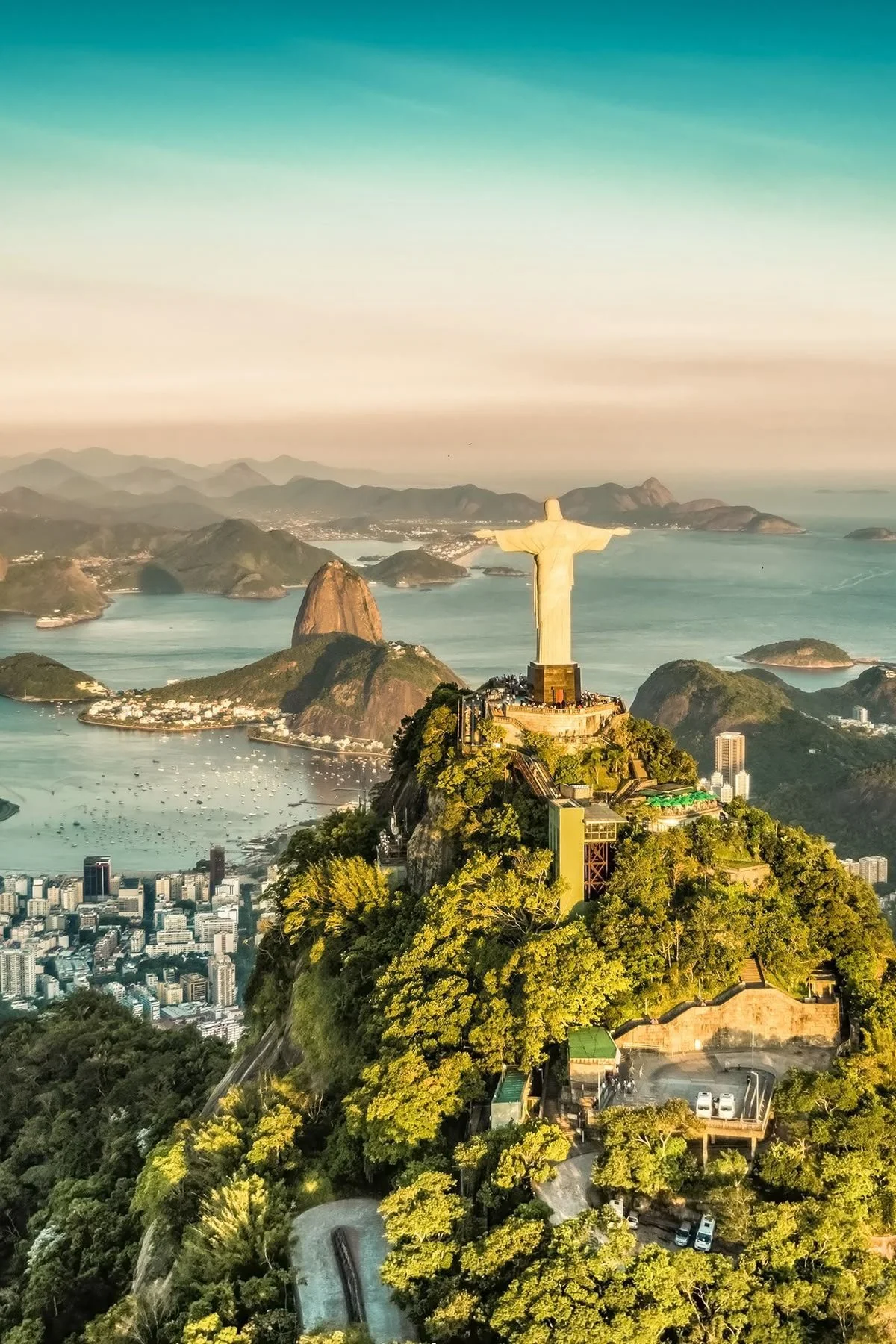 Aerial view of the Christ the Redeemer statue on Mount Corcovado overlooking Rio de Janeiro, Brazil, with mountains, waterways, and cityscape in the background at sunset.