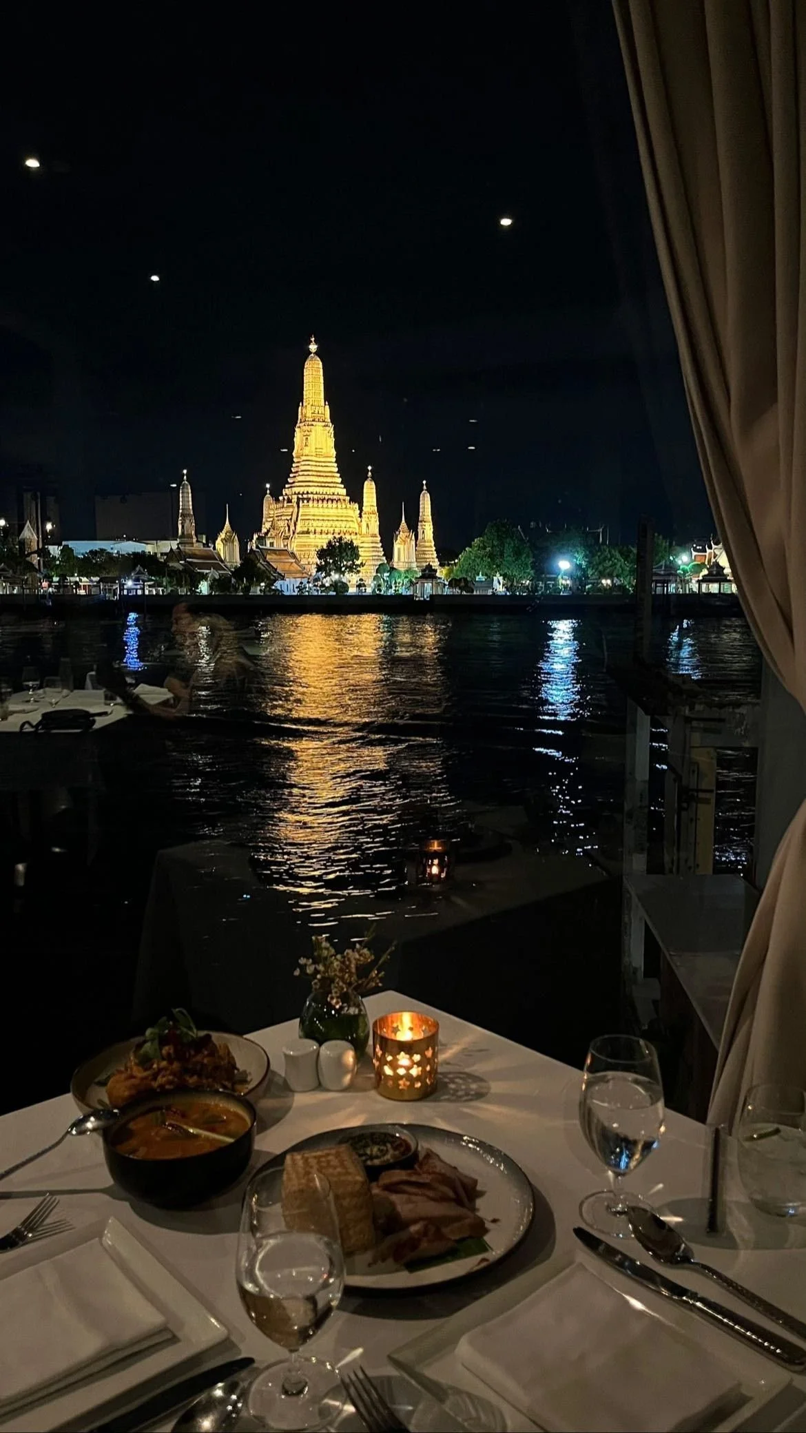 Night view of Wat Arun temple across the water, seen from a restaurant table with food, glasses, and candles.