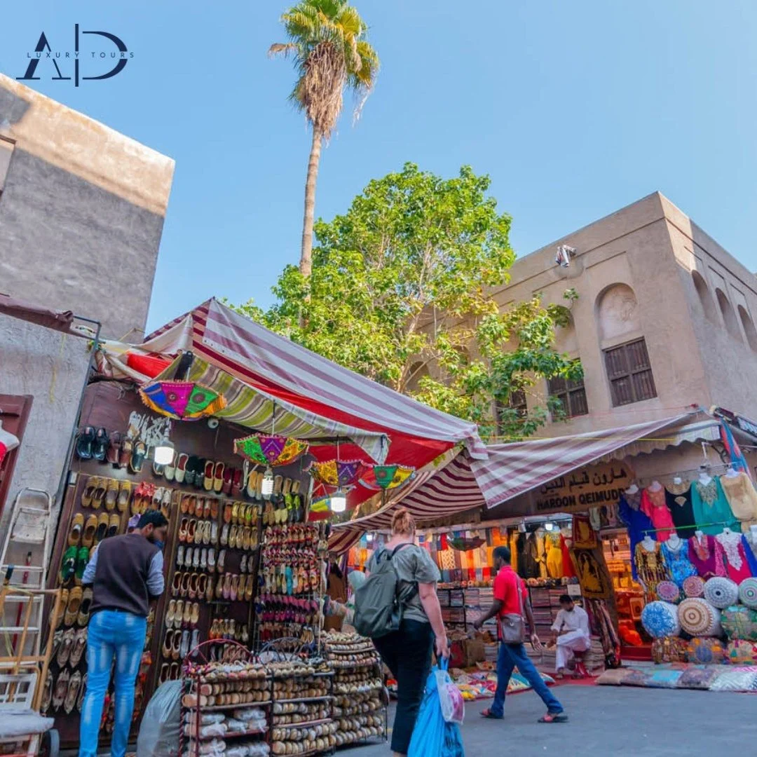 Outdoor market with stalls selling shoes, bags, and cushions. People browsing under striped awnings with colorful umbrellas. Tall palm trees and a building in the background. Clear blue sky.