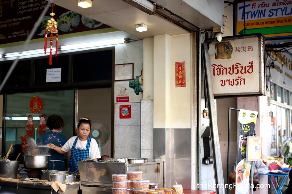 Street food stall with two women cooking and preparing food, in front of a shop with signs in multiple languages including Thai and English, decorated with red hanging ornaments.