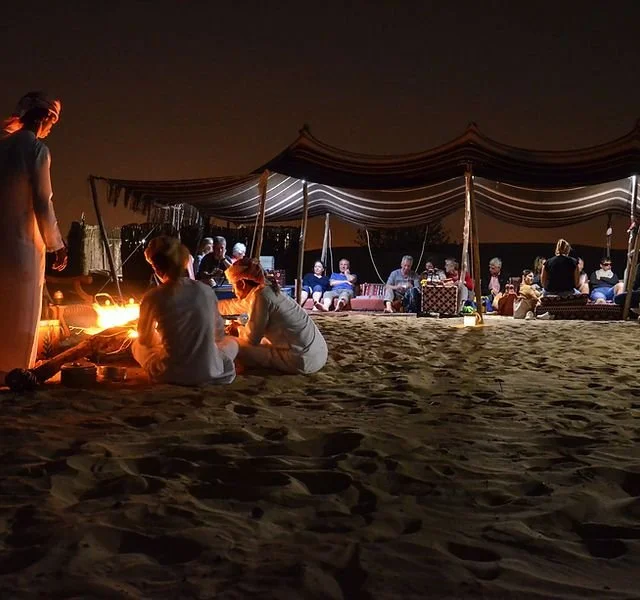People gathered around a bonfire on the beach at night with a large tent in the background.