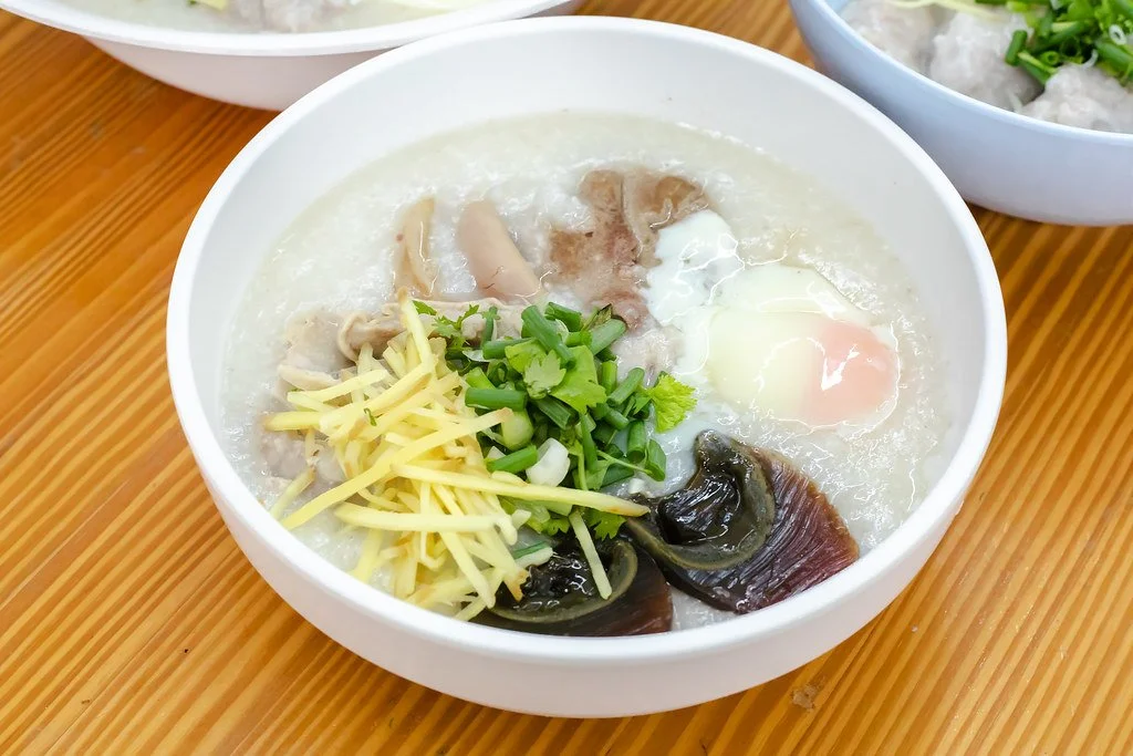 Bowl of congee with shredded cheese, chopped green onions, a poached egg, sliced chicken, and preserved eggs on a wooden table.
