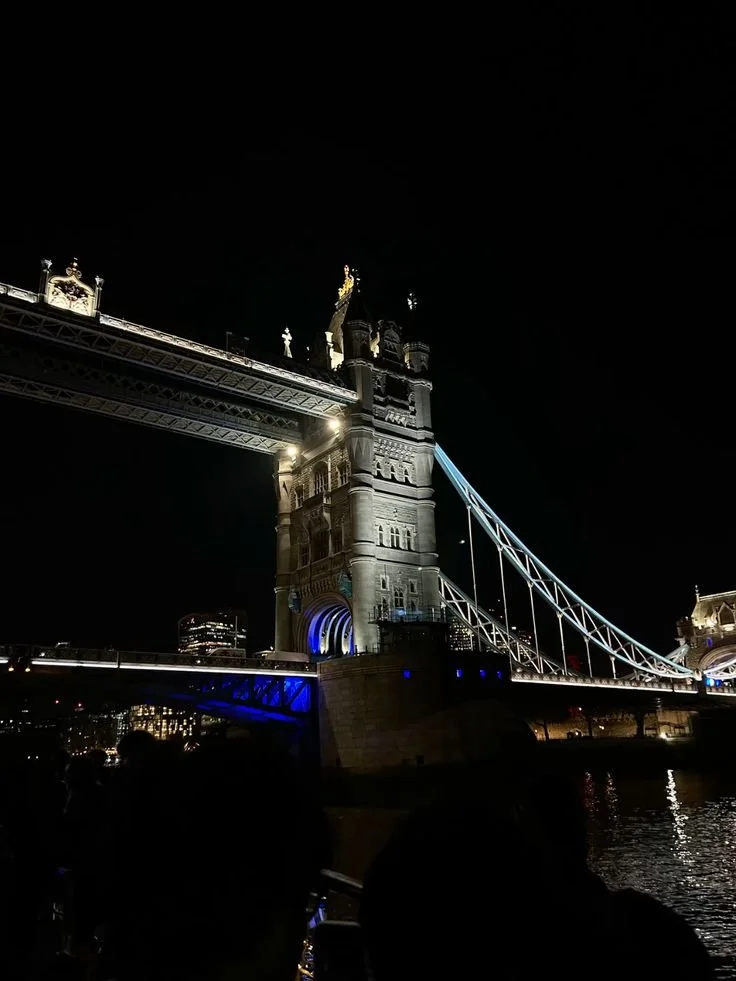 Night view of Tower Bridge in London, illuminated with lights, showing the bridge's towers and suspension chains over the Thames River.