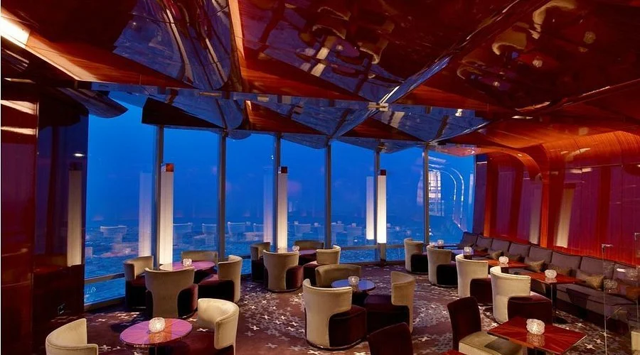 Upscale lounge or bar area with large panoramic windows, city skyline view at dusk, modern furniture, and ambient lighting.