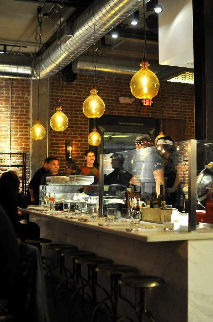 People inside a restaurant kitchen talking and working, with hanging gold light bulbs, exposed ductwork, brick wall, and a marble counter with glasses and plates.