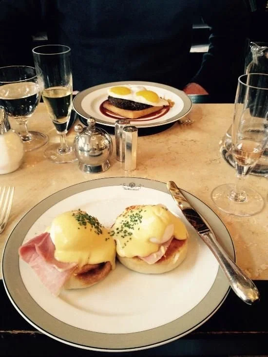 Two Eggs Benedict with ham and hollandaise sauce on a white plate, garnished with chopped chives, on a table set with glasses of water, champagne, and condiment shakers.