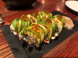 Sushi roll topped with green avocado slices, drizzled with dark eel sauce, served on a black rectangular platter with a small dish of soy sauce nearby.