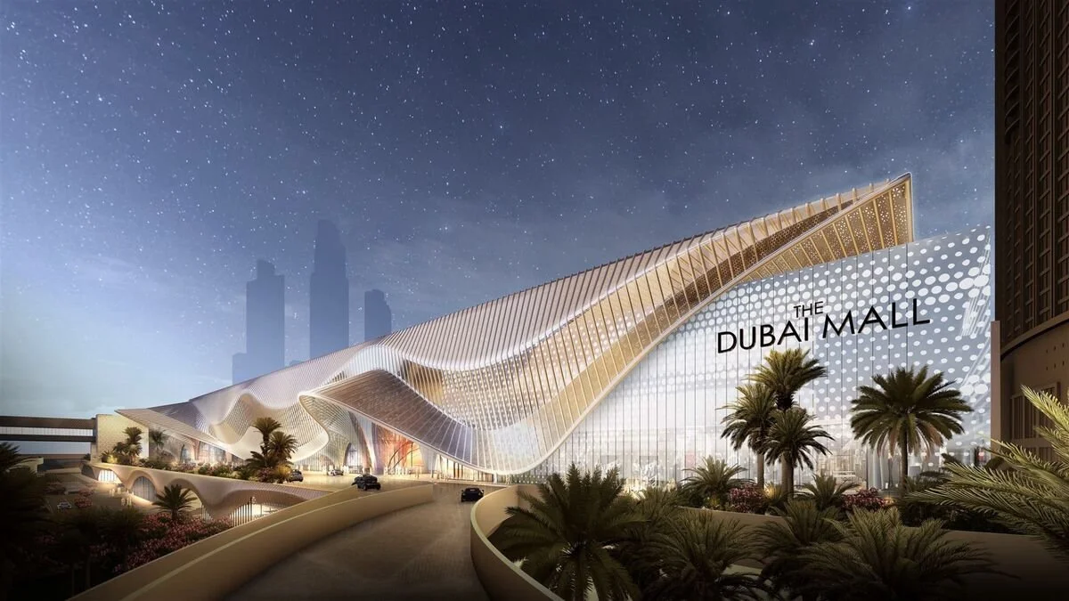 Rendering of Dubai Mall building at night with a wavy architectural design, palm trees in front, and Dubai skyscrapers in the background.