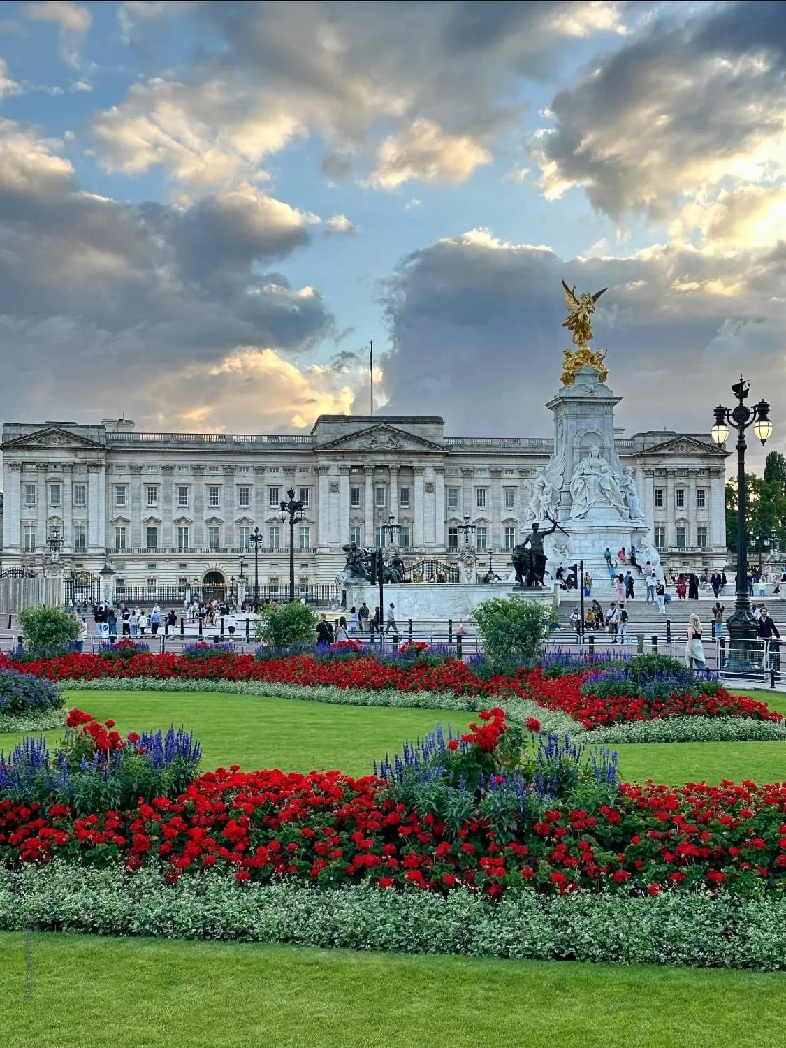 The image shows Buckingham Palace in London with a large garden in the foreground filled with colorful flowers, and a statue with a golden sculpture on top, under a sky with clouds.