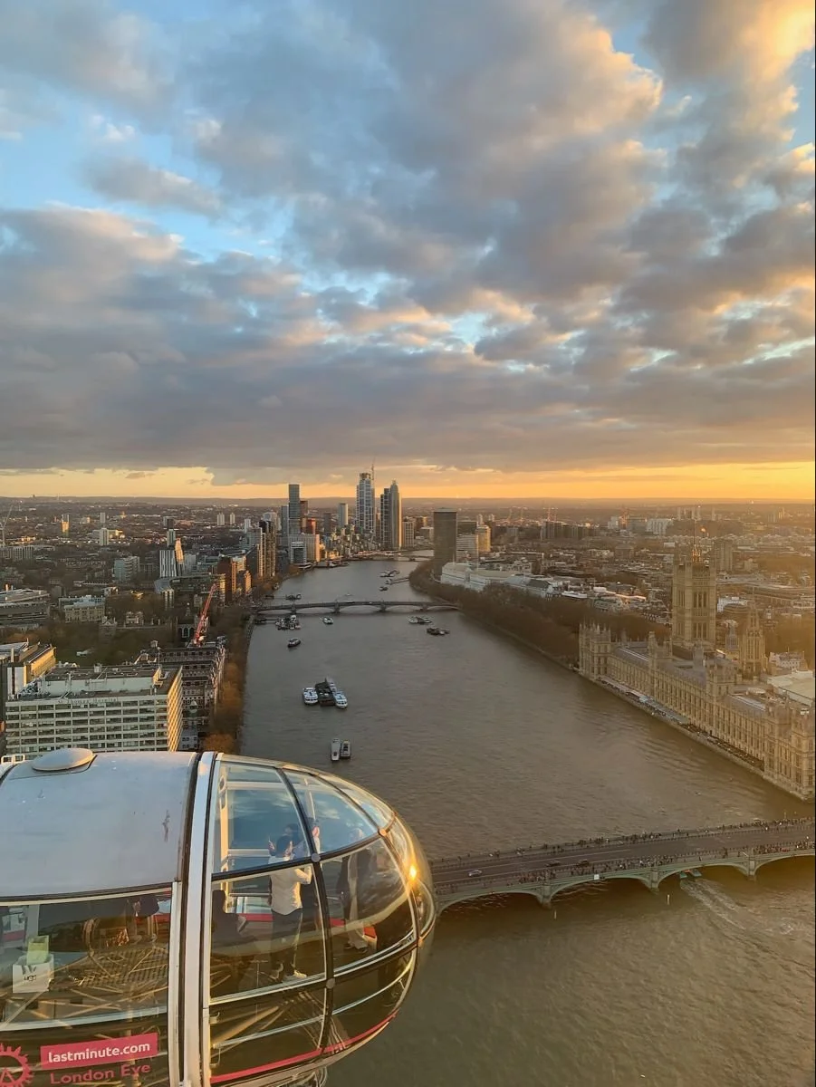 View of the London skyline at sunset with the River Thames, boats, and the London Eye in the foreground.