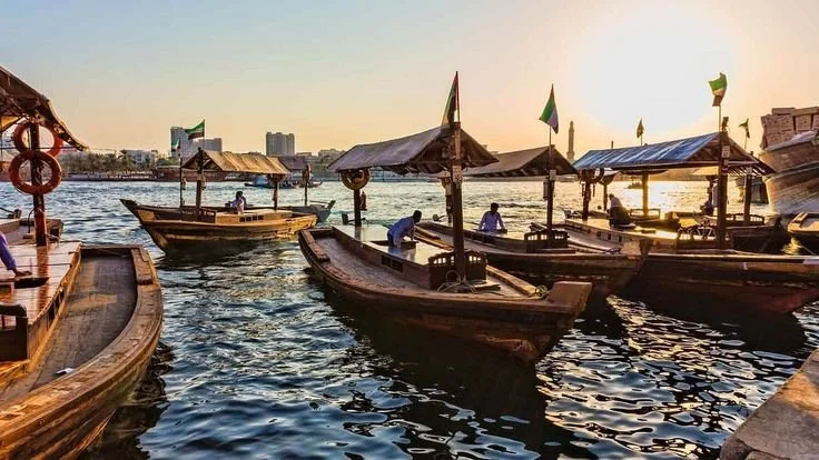 Several wooden boats docked on a river at sunset with city buildings in the background.
