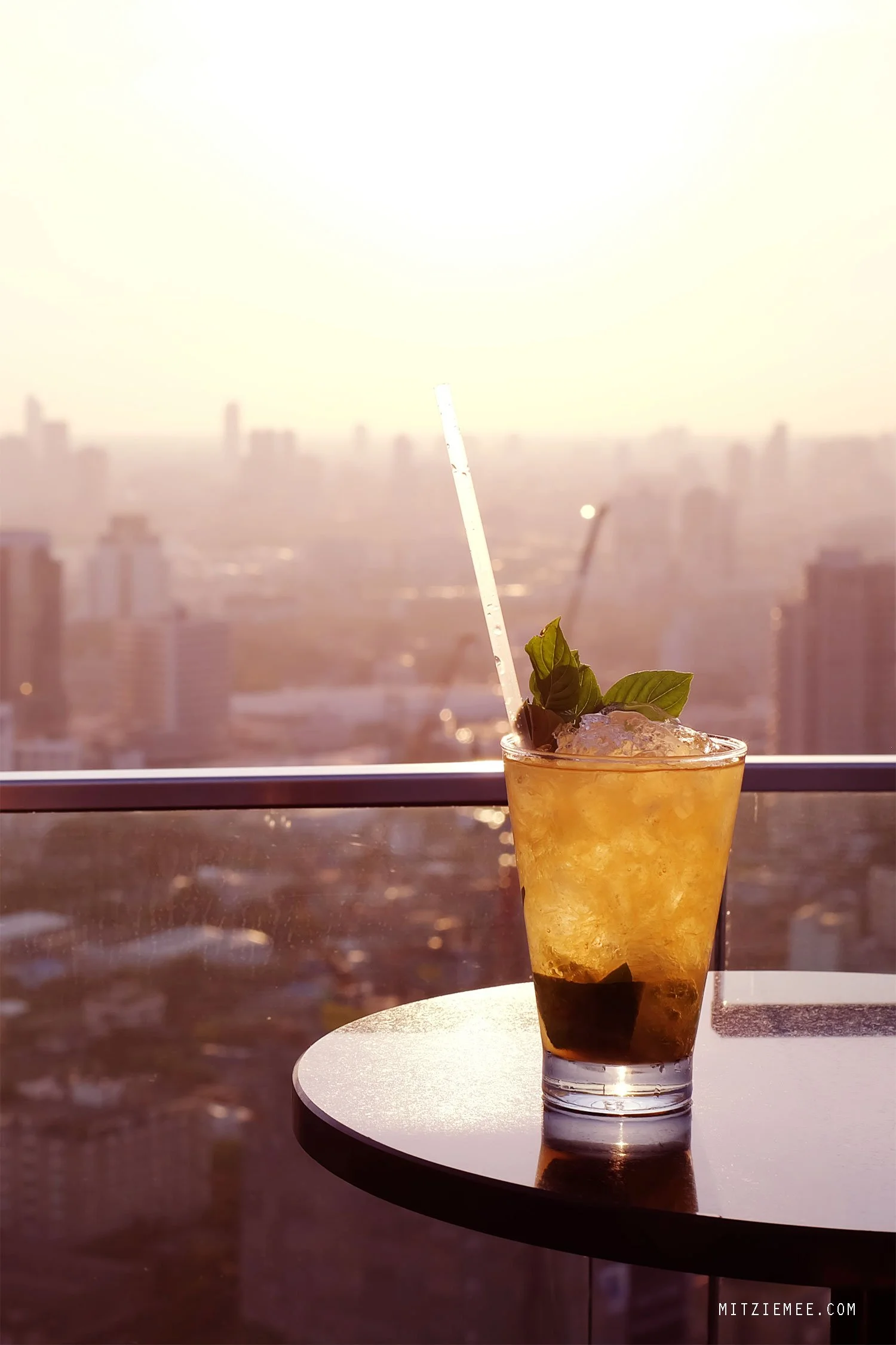 A glass of iced beverage garnished with mint leaves on a table, with a city skyline and sunset in the background.