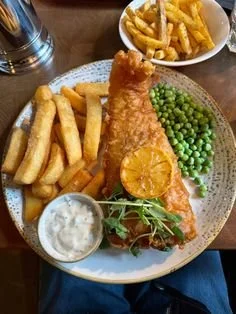 Plate of fish and chips with French fries, green peas, lemon wedge, and tartar sauce, along with a bowl of French fries in the background.