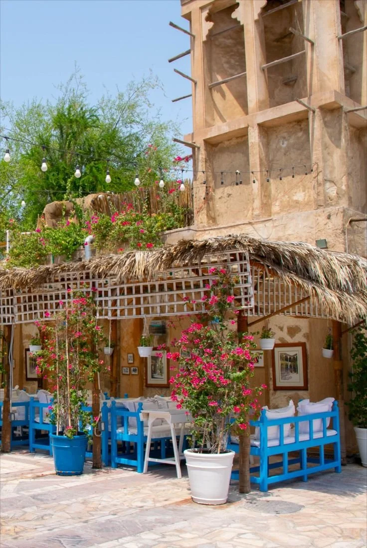Outdoor seating area with blue benches, white tables, pink flowering bushes in pots, and a thatched roof covering, adjacent to a rustic building with a tower and string lights.