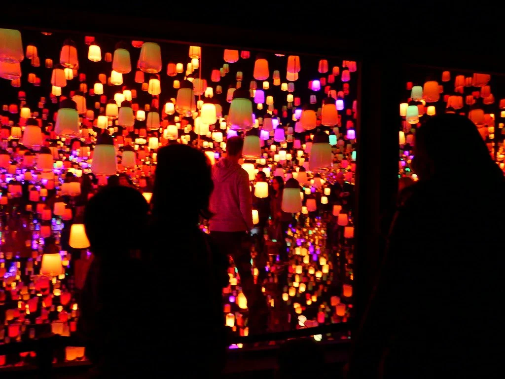 People viewing an art installation with hanging lanterns and colorful lights.