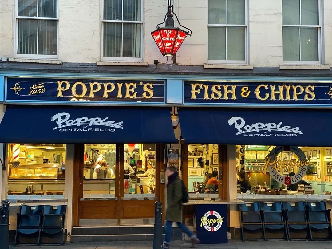 Front view of Poppie's Fish & Chips restaurant with blue awnings and signage, showing people inside and a pedestrian walking by outside.