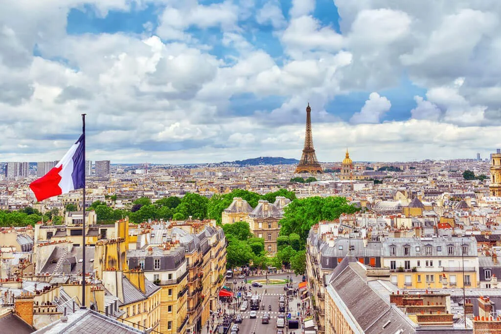 View of Paris cityscape with the Eiffel Tower in the distance, French flag flying in the foreground, atop buildings with classic Parisian rooftops, green trees, and a cloudy sky.