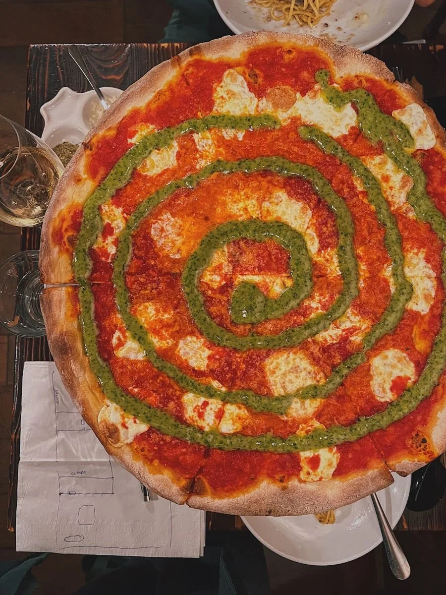 A large pepperoni pizza with green sauce in a spiral pattern on top, served on a wooden table with drinks and side dishes around it.