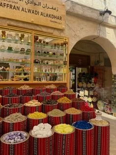 A street market stall with colorful spices in bowls and shelves of small bottles on a glass display case above, located outside a building with an arched entrance and a sign in Arabic and English.