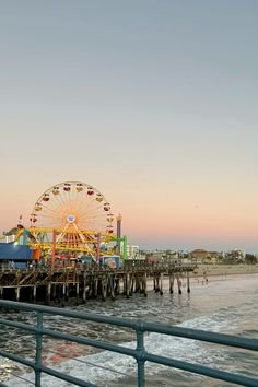 View of a pier with a Ferris wheel, amusement rides, and structures at sunset, over the ocean.