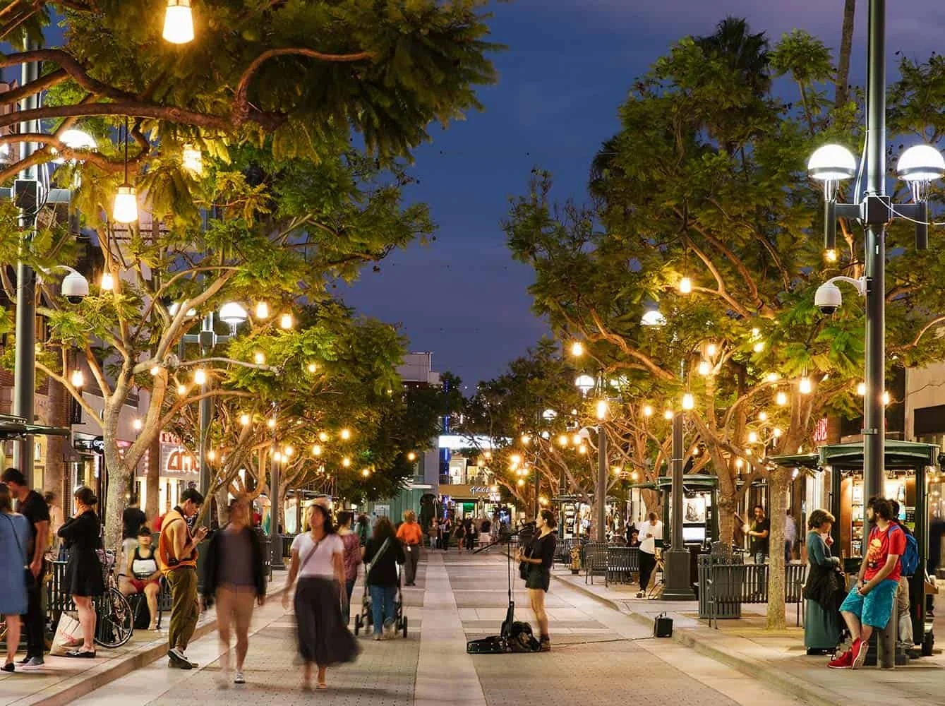 People walking and chatting on a lively street with trees, decorative hanging lights, and street lamps during evening.