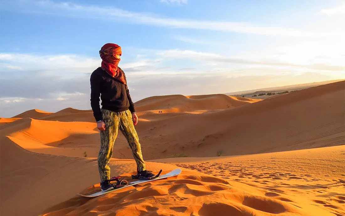 Person on a snowboard standing on sand dunes in a desert at sunset, wearing a black hoodie, patterned pants, and a red scarf over their head.