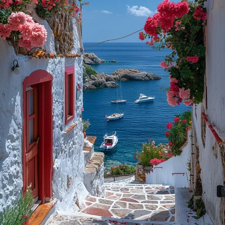 A seaside village view with white-washed buildings, red window frames, and vibrant pink flowers, overlooking a calm blue sea with sailboats anchored nearby.