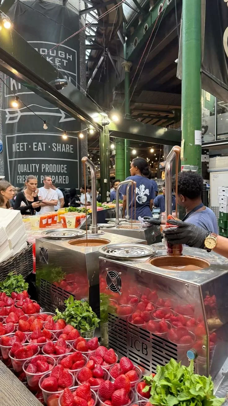 A bustling market stall with fresh strawberries, green herbs, and chocolate fountains surrounded by people, inside a modern market with high ceilings and string lights.
