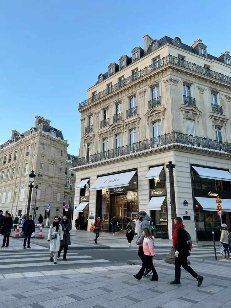 A busy city street in front of a Louis Vuitton store, with pedestrians crossing and walking past on a clear day.