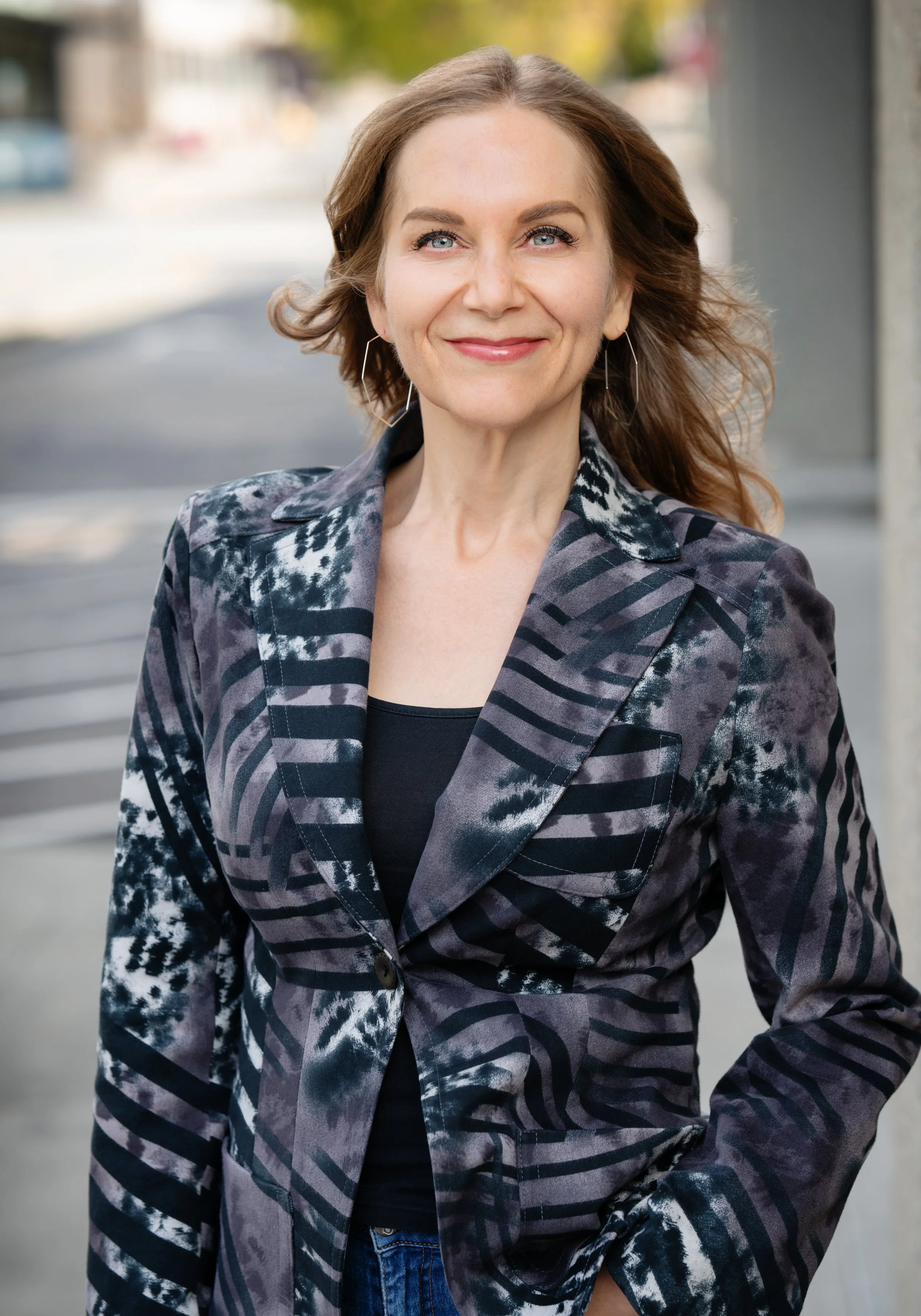 Susan Hovey, Founder of SMASH Marketing & Success Coaching, with light skin, long light brown hair, and blue eyes wearing a patterned blazer and earrings, standing outdoors on a city street with blurred background.