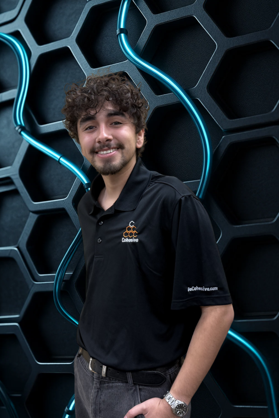 A young man with curly hair and a beard smiling, wearing a black shirt with a company logo and website, standing in front of a black honeycomb-patterned wall with blue neon lights.