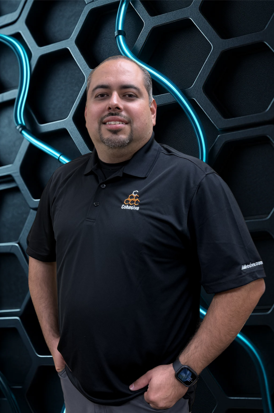 A man wearing a black Polo shirt with the company logo 'Cohesive' on it, standing in front of a hexagonal patterned wall with neon blue light accents.
