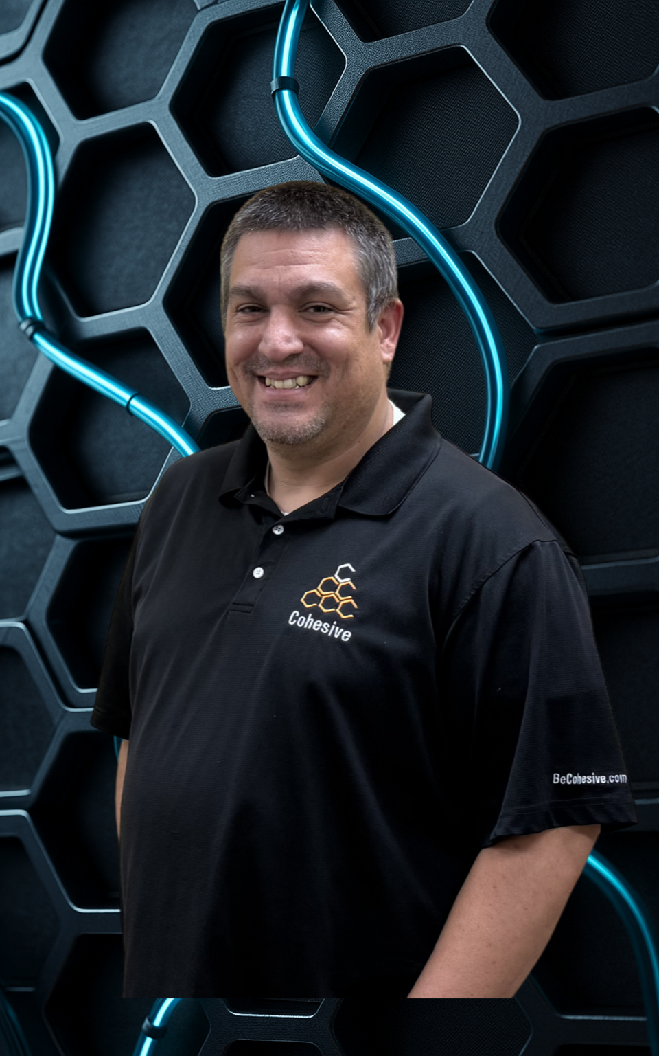 A man with short dark hair and a beard smiling, wearing a black polo shirt with a logo and the word 'Cohesive' on it, standing in front of a hexagonal patterned background with blue neon tubing.