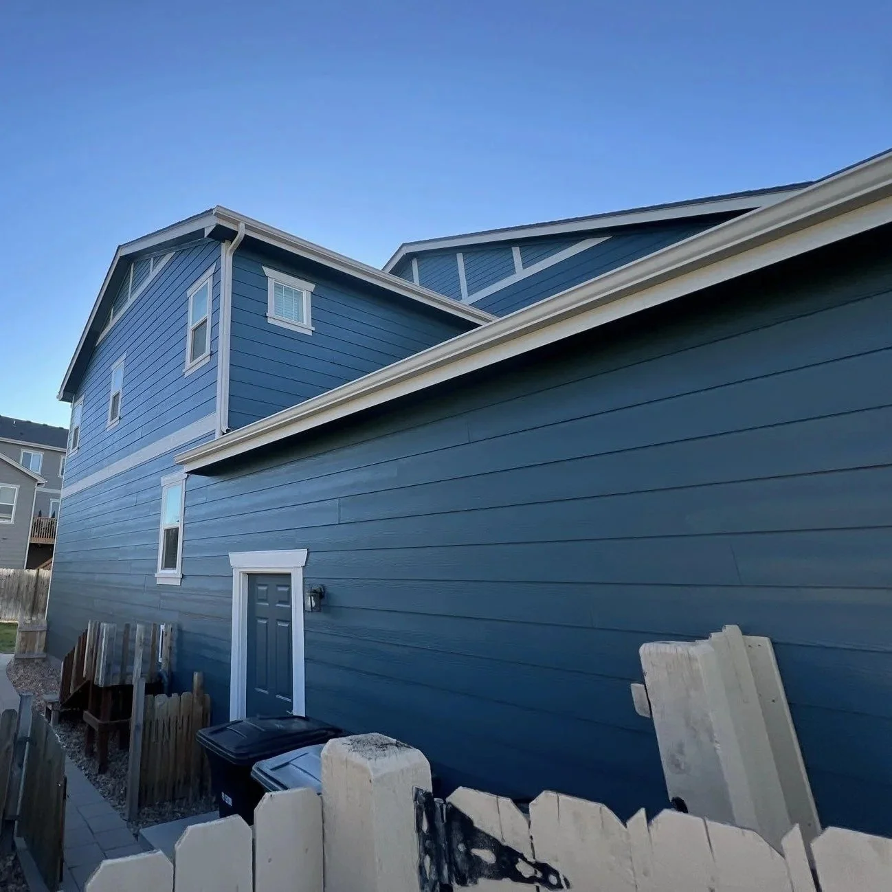 A blue house with white trim, partly visible with multiple stories and sloped roofs; a small fenced yard with a trash can and building materials nearby; clear blue sky.