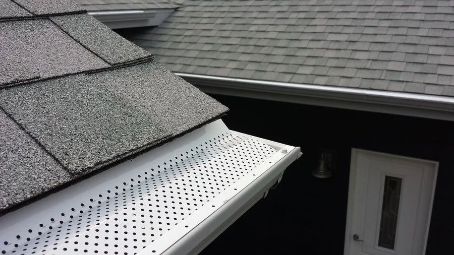 Close-up of a house roof with asphalt shingles and a white perforated gutter system.