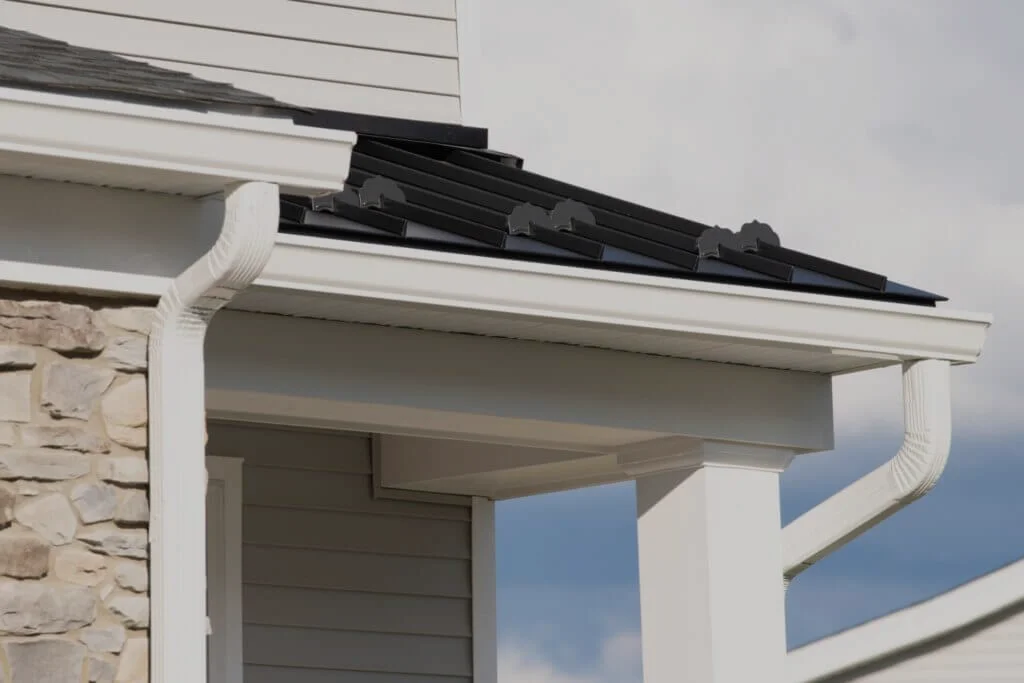 White seamless gutters and downspouts installed below a black metal roof on a stone-and-siding home.