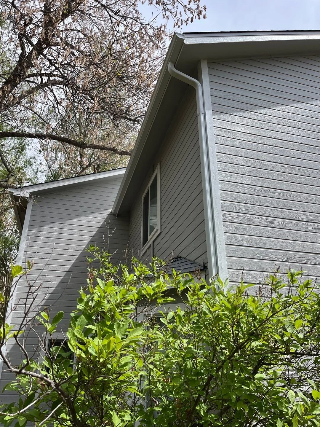 Seamless gutter and downspout installation completed along the side of a light-colored home with mature landscaping.