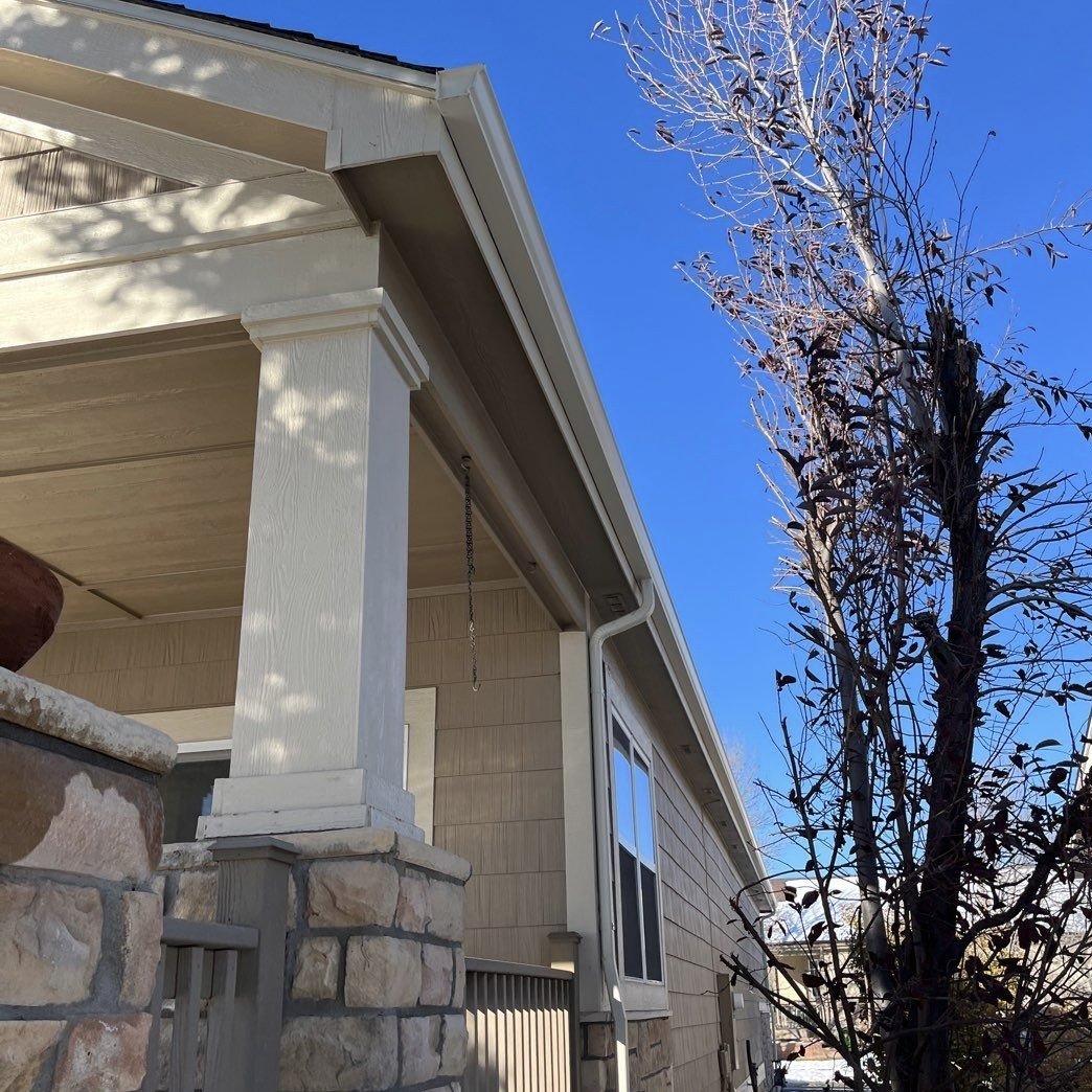 Close-up of a house porch with beige siding, white trim, and a stone pillar, next to a leafless tree under a bright blue sky.