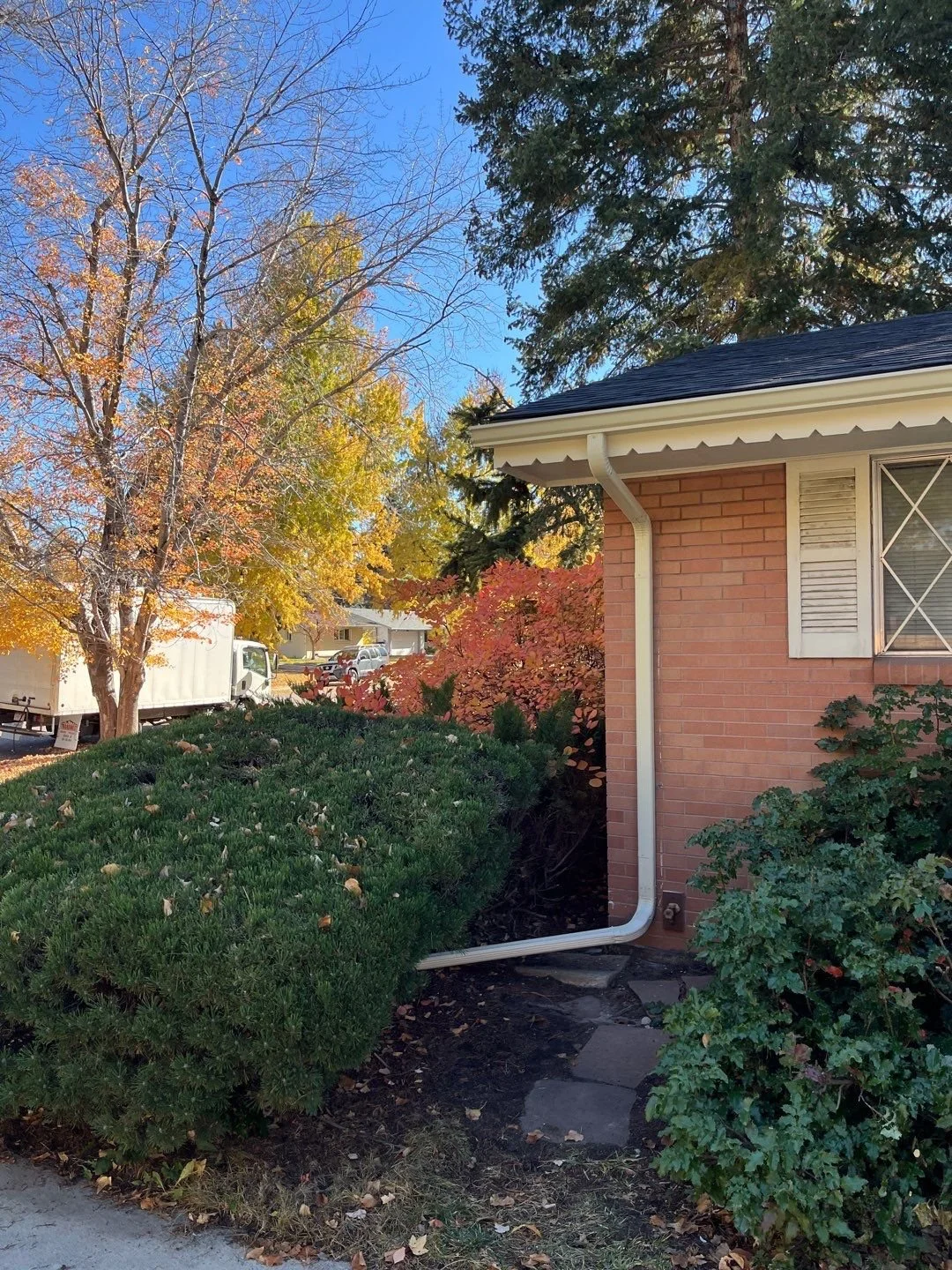 Seamless gutters and downspouts installed on a brick home surrounded by mature landscaping.