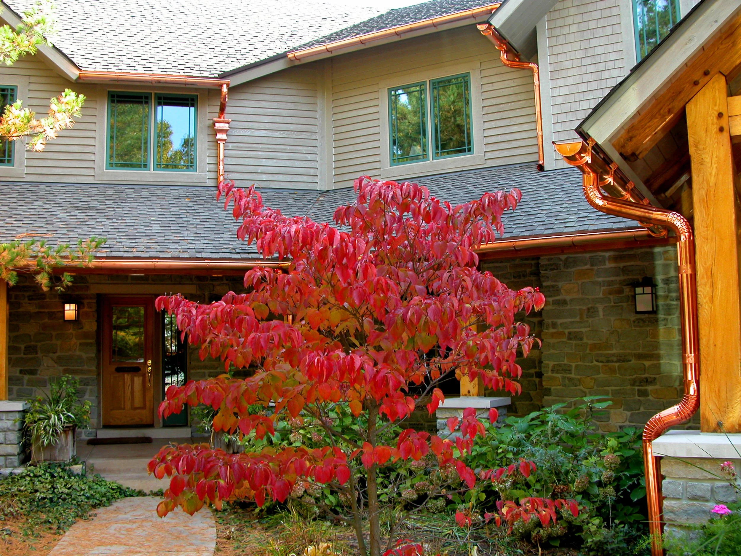 Copper half-round gutters and round downspouts installed across the multiple rooflines of a stone-and-siding home.