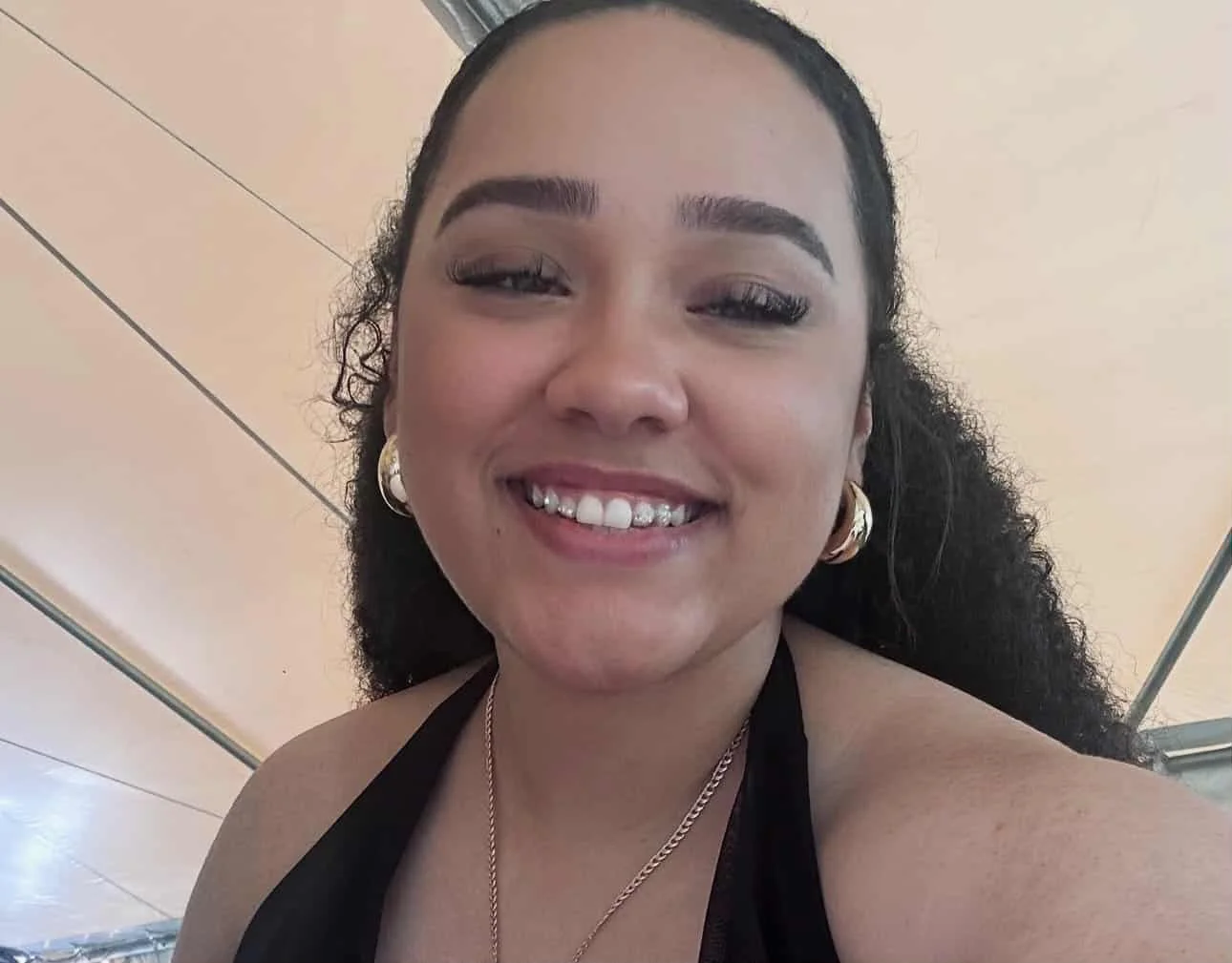 A woman smiling with braces, wearing gold hoop earrings and a black top, in an indoor setting with beige walls.