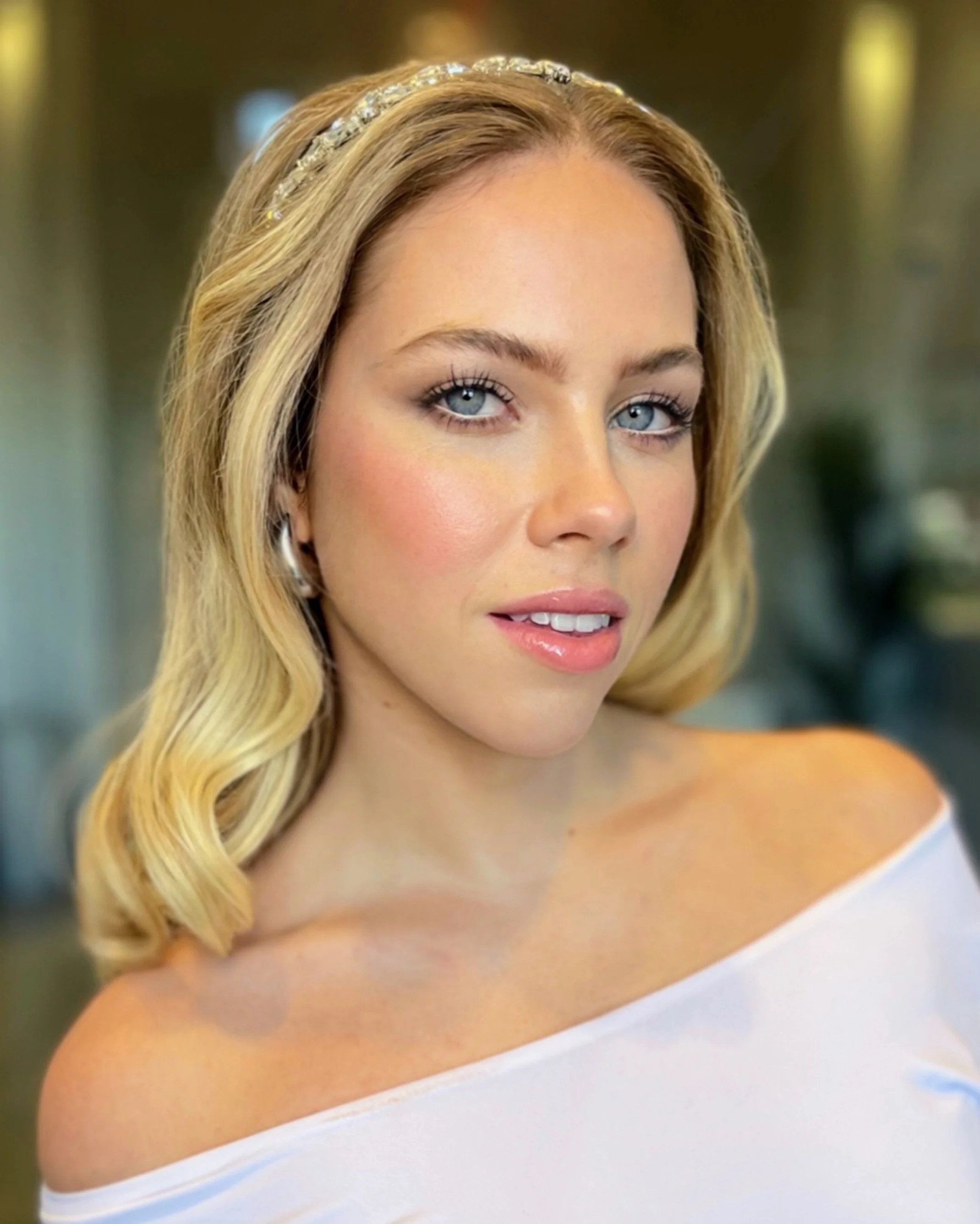 Close-up of a woman with blonde wavy hair, decorated with a jeweled headband, wearing a strapless white dress.