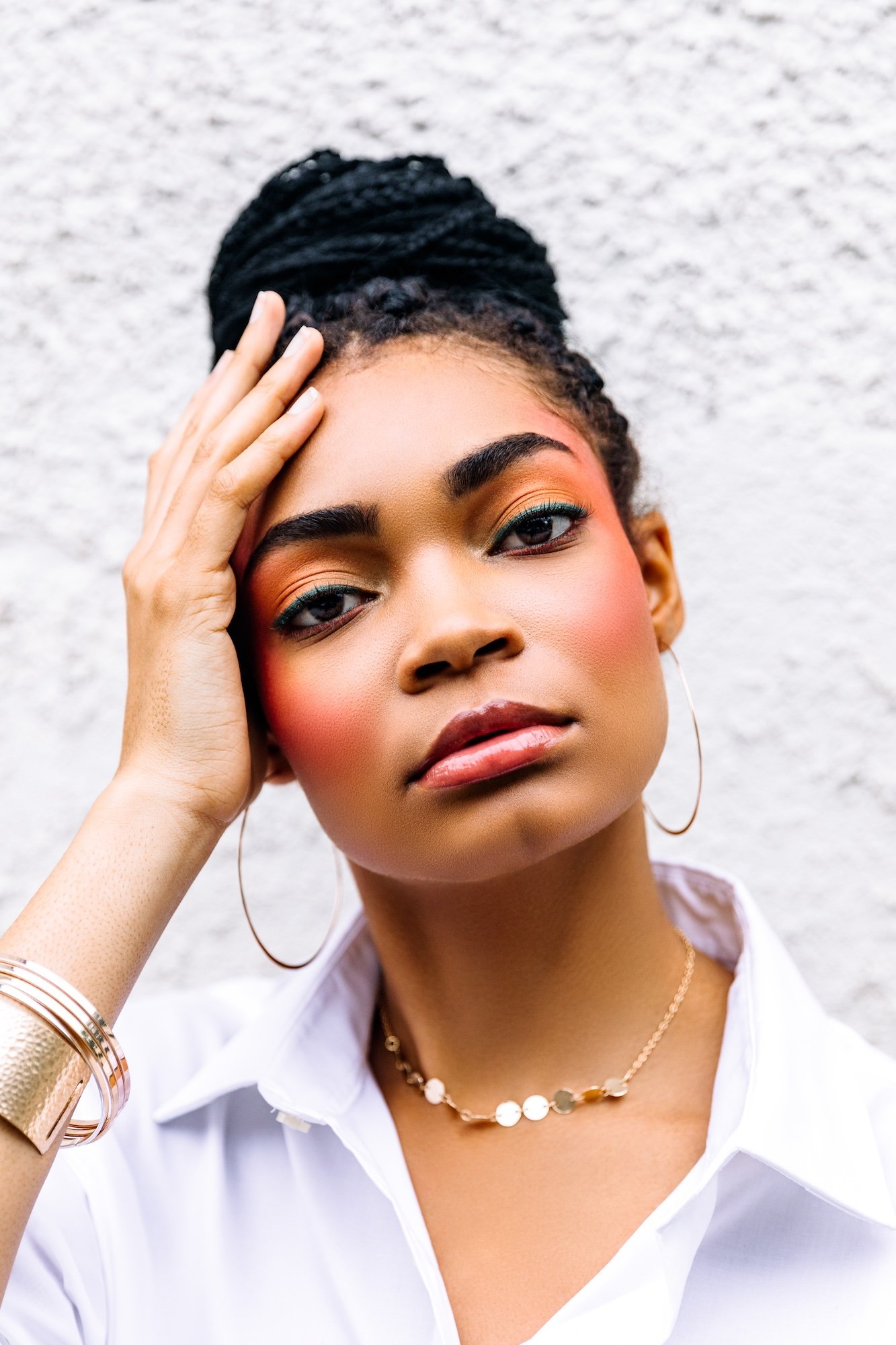 A woman with dark braided hair styled in a bun, wearing hoop earrings, a gold necklace, and a white shirt, posing against a textured white wall.