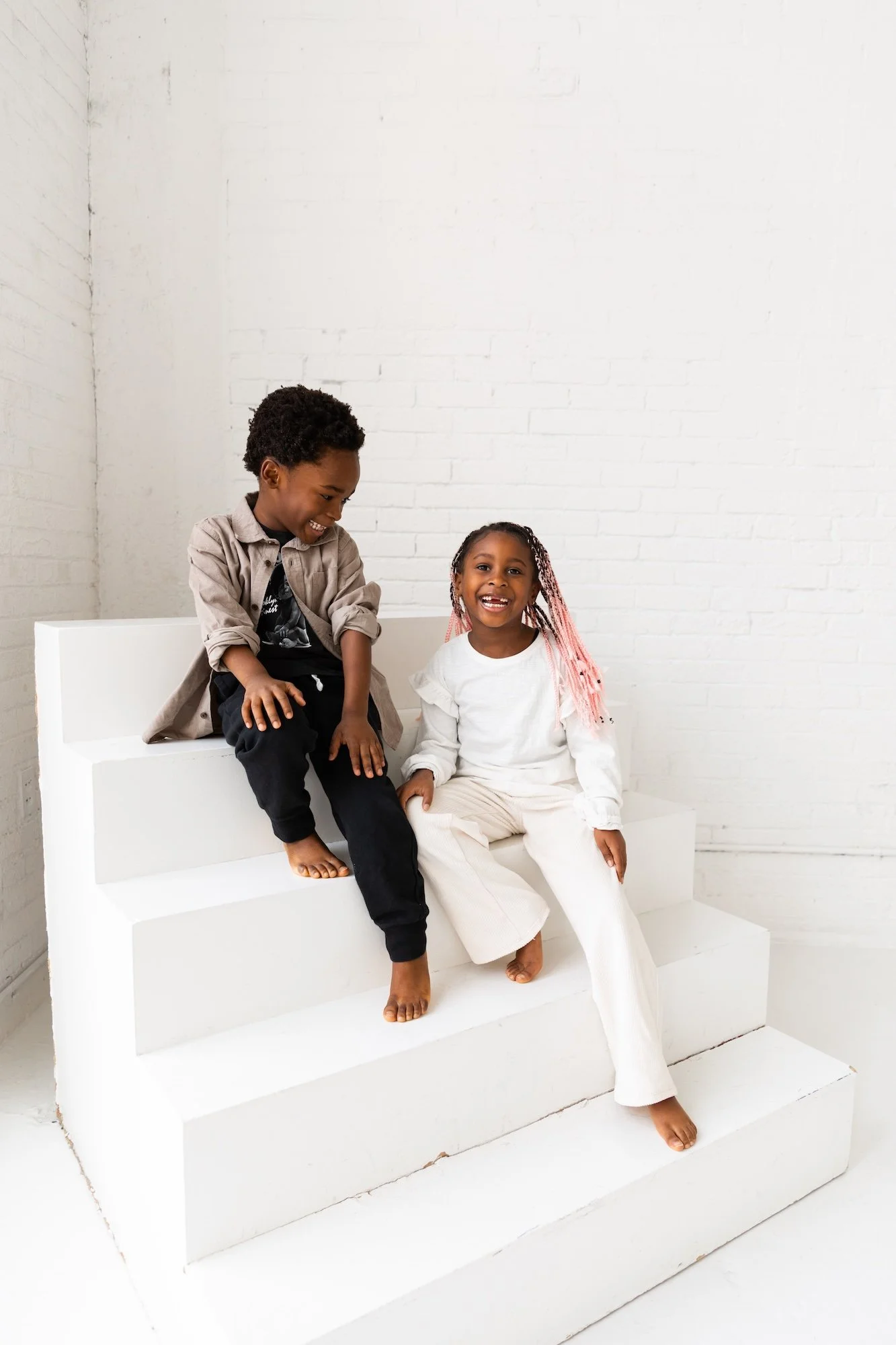 Two children, a boy and a girl, sitting on white stairs in front of a white brick wall, smiling and laughing together.