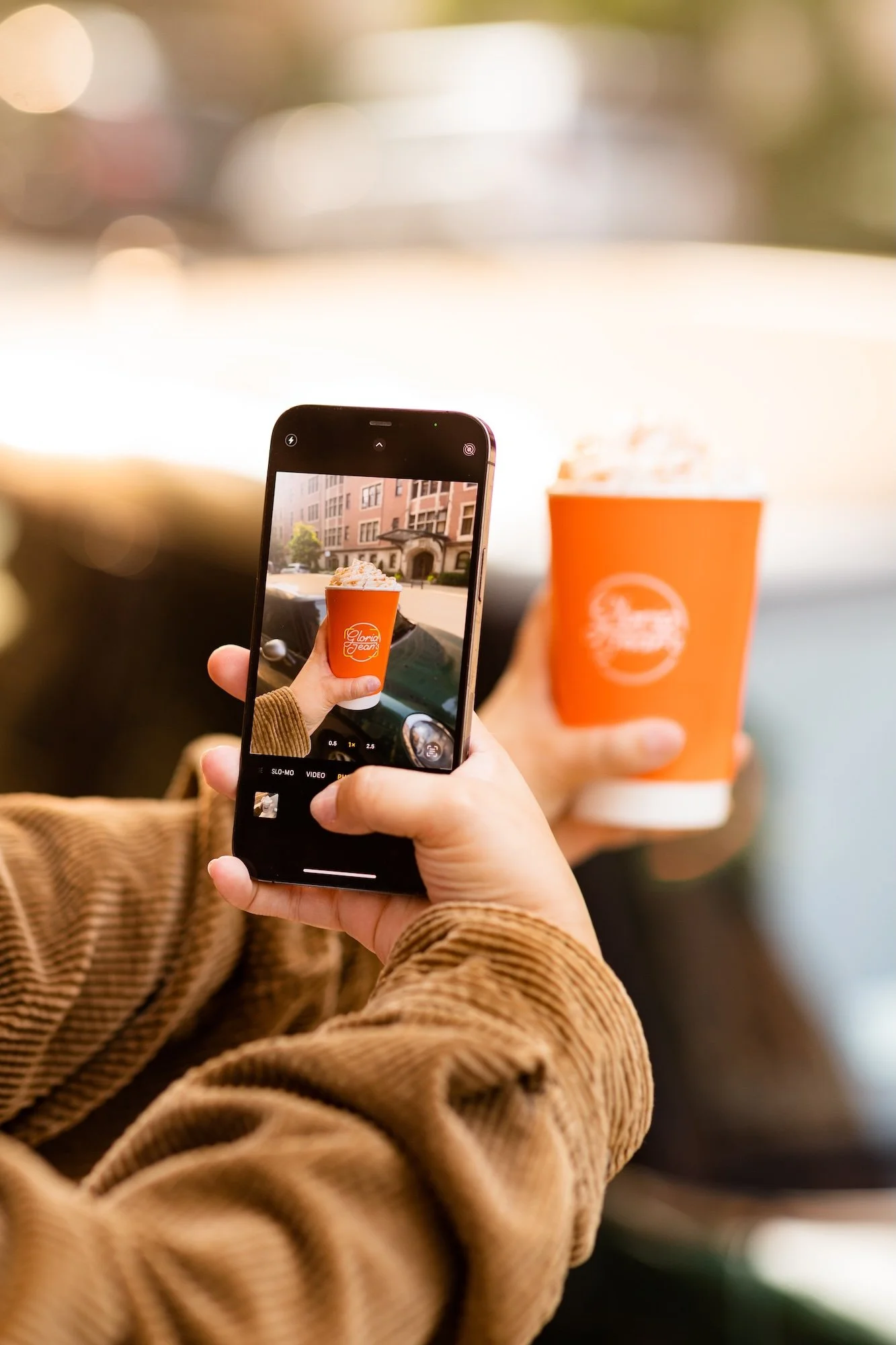 Person taking a photo of a bright orange cup with whipped cream, showing the cup's logo, inside a vehicle.